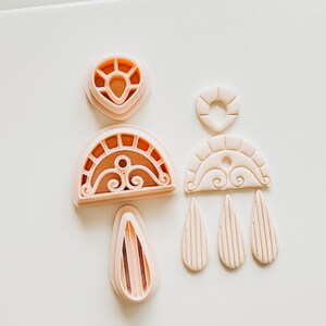 May include: A selection of light pink polymer clay earring cutters in various shapes. The cutters include teardrops, arches, and circular designs, ideal for crafting handmade jewellery. The shapes are arranged on a white surface.