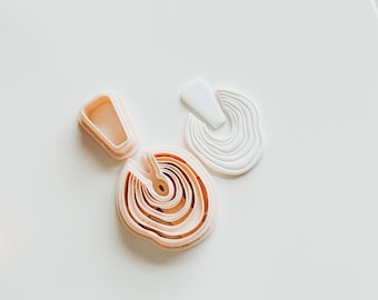 May include: A set of two cookie cutters in a light pink color. The larger cutter is a circle with concentric rings, while the smaller cutter is a crescent shape.