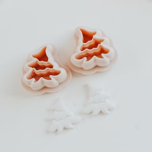 Könnte beinhalten: Zwei Ausstecher in Weihnachtsbaumform mit hellrosa Außen- und orangefarbenem Innenbereich sowie zwei weiße, baumförmige Kekse. Die Ausstecher haben einen gewellten Rand. Die Artikel befinden sich auf einer weißen Oberfläche.