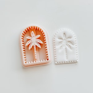 May include: Two pink plastic cookie cutters, one with a palm tree design and the other with a raised palm tree design.