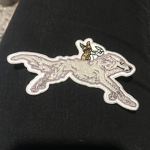 以下が含まれることがあります： 狼が走っていて、その背中に小さな猫と鳥が乗っている、白い刺繍のワッペンです。