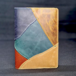 May include: A multi-coloured leather notebook cover with a patchwork design. The cover is made of four different colours of leather: brown, tan, green, and purple. The seams are visible and stitched with a dark thread.