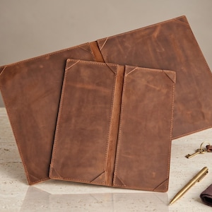 May include: Three brown leather desk accessories: a large desk mat, a smaller notepad, and a bi-fold wallet. The leather has a distressed finish with stitched corner details. A gold pen, keys, and a small brown cloth are also visible.