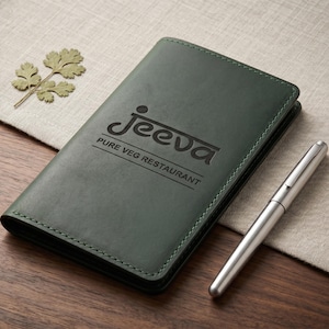 May include: A dark green leather menu cover with the text "Jeeva Pure Veg Restaurant" embossed on the front. A silver pen rests beside the menu on a wooden surface. A sprig of dried leaves is also present.