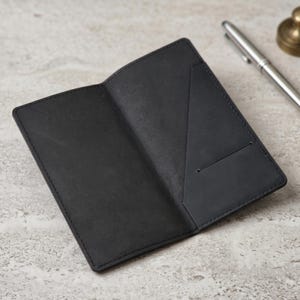 May include: An open, black leather passport holder with a pocket on the right side. A silver pen and a small brass paperweight are in the background on a light gray surface.