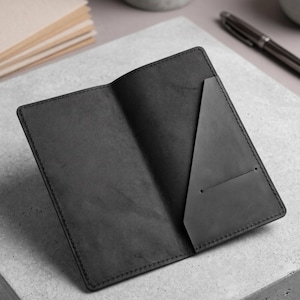 May include: Open black leather wallet with a pocket on the right side. The wallet is on a gray surface, with a stack of beige notepaper and a pen in the background. The wallet is open, revealing the interior.
