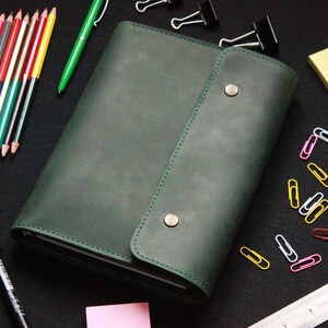 Peut inclure: Un carnet en cuir vert foncé avec deux boutons pression argentés et une bordure cousue. Le carnet est entouré de divers fournitures de bureau, y compris des crayons, un surligneur, un marqueur noir, des trombones et une règle.