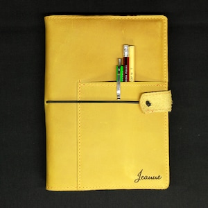 May include: A yellow leather bound journal with a black elastic closure and a pocket on the front cover. The journal has the name "Jeanne" embossed on the front cover. There are several pens and pencils in the pocket.