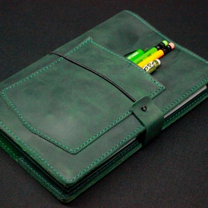 May include: A dark green leather journal with a flap closure and a pocket for pencils. The journal has a strap closure and is stitched with white thread.
