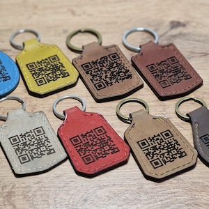 May include: Eight leather keychains with different colored leather and a black and white QR code design. The keychains are on a wooden surface.
