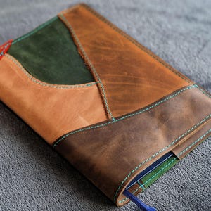 May include: A handcrafted leather journal cover with a patchwork design. The cover features various shades of brown, green, and red leather, stitched together with teal thread. A blue ribbon bookmark is visible.
