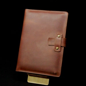 May include: A brown leather journal with a strap closure and brass accents. The journal is closed and sitting on a wooden stand.