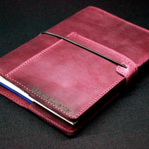 May include: A burgundy leather bound journal with a black elastic closure and a pocket on the front cover. The journal has the text "Filofax" embossed on the front cover.