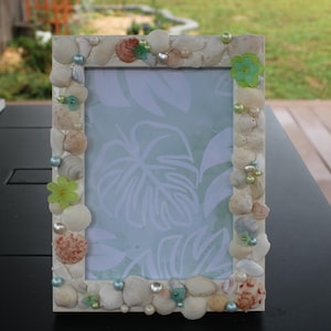 Op de afbeelding: Een rechthoekige fotolijst versierd met schelpen, parels en kleine groene en blauwe accenten. De lijst heeft een lichtgekleurde achtergrond en een lichtblauw en wit patroon aan de binnenkant. De lijst staat op een zwart oppervlak.