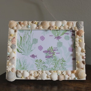 May include: A rectangular photo frame decorated with various seashells, pearls, and small beads. The frame surrounds a picture with an underwater theme, featuring purple fish and green seaweed. The frame is approximately 8 inches wide and 6 inches tall.