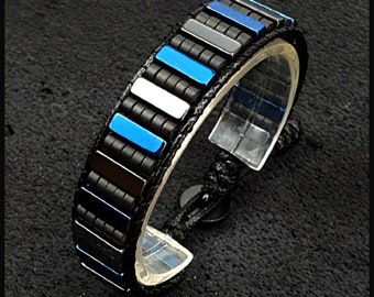 Può includere: Un braccialetto nero con perline rettangolari blu, argento e nere. Il braccialetto è realizzato con un cordoncino intrecciato e ha una chiusura a nodo scorrevole.