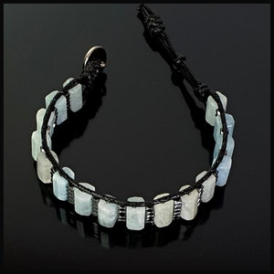 K&ouml;nnte beinhalten: Ein Armband aus hellblauen Aquamarin-Steinen und schwarzem Band. Die Steine sind rechteckig und haben eine leicht unregelm&auml;&szlig;ige Form. Das Band ist am Ende mit einem Knoten gebunden.
