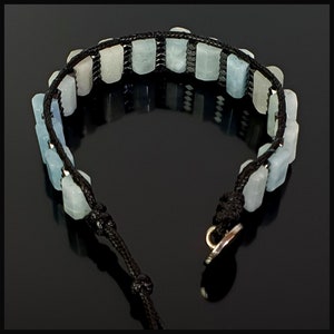 K&ouml;nnte beinhalten: Ein Armband aus schwarzem Band mit hellblauen rechteckigen Perlen. Das Armband hat einen silbernen Verschluss.