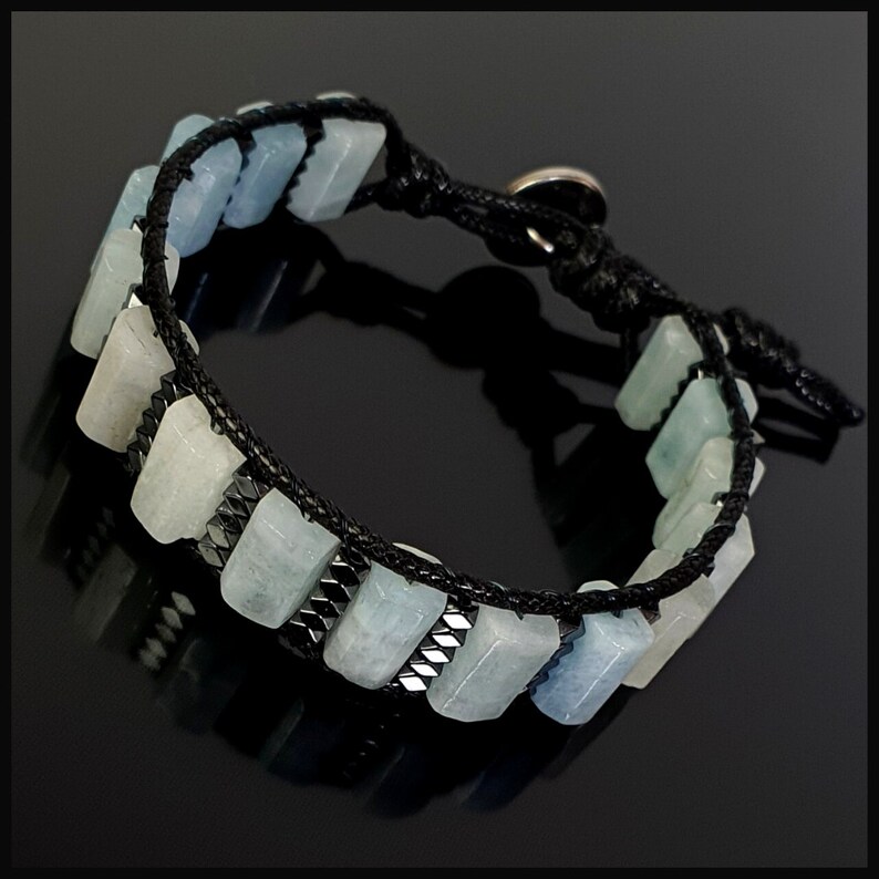 K&ouml;nnte beinhalten: Ein Armband aus schwarzem Band mit rechteckigen Perlen in Hellblau und Grau. Das Armband hat einen silbernen Verschluss.
