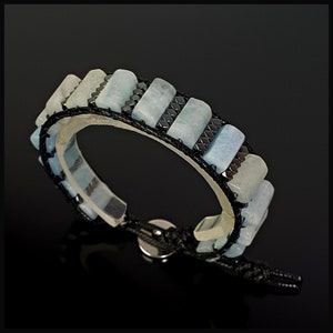 K&ouml;nnte beinhalten: Ein Armband aus schwarzem Band mit rechteckigen Perlen in Hellblau und Dunkelgrau. Das Armband hat einen Magnetverschluss.