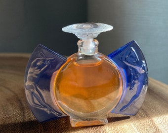 May include: A vintage perfume bottle with a clear glass body filled with amber liquid. The bottle has a clear, decorative stopper and is flanked by two blue, wing-shaped glass panels, each featuring a sculpted figure.