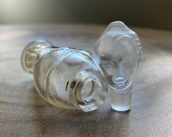 May include: A clear glass perfume bottle with a stopper shaped like a woman's face. The bottle is lying on its side, and the stopper is upright. The glass has a frosted appearance, and the details of the face are finely etched.