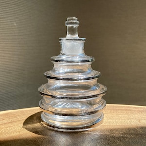 May include: A clear glass bottle with a stopper. The bottle has a tiered design with black trim. The stopper is frosted glass. The bottle is sitting on a wooden surface.