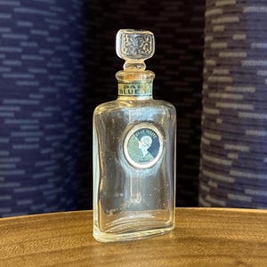 May include: A clear glass perfume bottle with a decorative stopper. The bottle has a rectangular shape with a label featuring a portrait and the words "Pale Blue". The stopper is square with an ornate design.