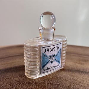 May include: A vintage perfume bottle with a clear glass body and a round stopper. The label reads "JASMIN" above "Andre Chenier" on a blue and white background. The bottle has a ribbed design on the sides.