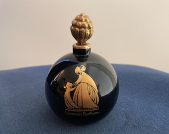 May include: A round, dark blue perfume bottle with a gold-colored, pinecone-shaped stopper. The bottle features a gold illustration of a woman and child, with the words "Lanvin Parfums" below.
