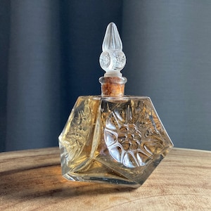 May include: A vintage amber glass perfume bottle with a decorative stopper. The bottle has a faceted design and embossed floral patterns. The clear stopper is pointed, and the bottle is sealed with a cork.
