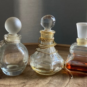 May include: Three antique perfume bottles with decorative stoppers. The bottles are clear glass, with one having a frosted ball stopper, another with a clear ball stopper, and the third with a white stopper. One bottle has a gold chain around the neck.
