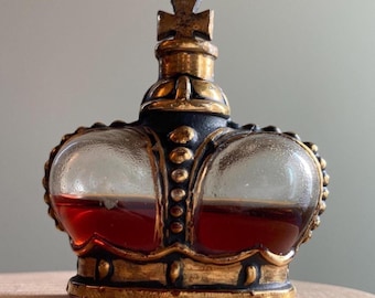 May include: A decorative glass bottle shaped like a crown, filled with a reddish-brown liquid. The bottle has gold-colored accents and a cross on top. The crown-shaped bottle is a unique decorative item.