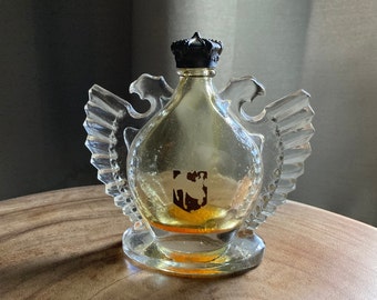 May include: A vintage perfume bottle with a clear glass body and a black crown-shaped stopper. The bottle has clear, wing-like accents on either side and a base. A small emblem is visible on the front of the bottle.