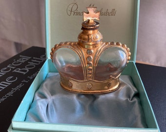 May include: A vintage crown-shaped perfume bottle with a gold-colored metal frame and a clear glass body. The bottle is inside a light blue box with a satin lining. The box has the words "Prince Matchabelli" printed on the inside.