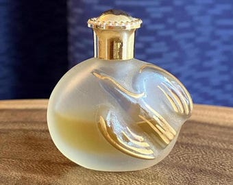 May include: A small, round perfume bottle with a frosted glass finish. The bottle features gold-colored accents in a bird-like design and a gold-colored cap. The bottle contains a yellow liquid.