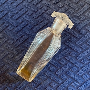 May include: A clear glass perfume bottle with a faceted, elongated body and a matching geometric glass stopper. The bottle features embossed floral detailing along the sides and contains a small amount of amber-coloured liquid at the base.