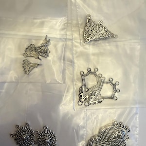 May include: A collection of silver-toned metal jewelry findings, including ornate filigree pendants and connectors. The pieces are in various shapes, such as sunbursts, triangles, and leaf-like designs, and are displayed in clear plastic bags.