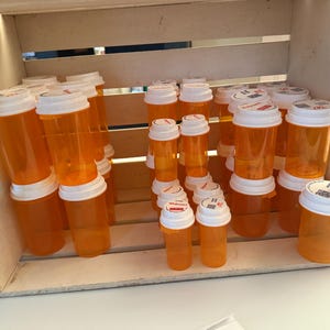 May include: A collection of empty orange plastic prescription pill bottles with white caps, arranged in various sizes within a light-colored wooden crate. The bottles feature white lids and some have small printed labels on the top surfaces.