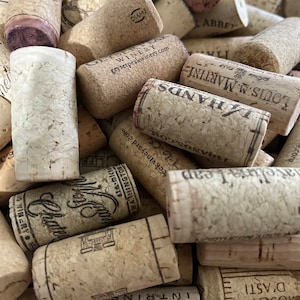 May include: A collection of used wine corks, featuring various brands and winery names. The corks are primarily beige and brown, with some showing remnants of red wine. Some corks have printed text, including winery names and logos.