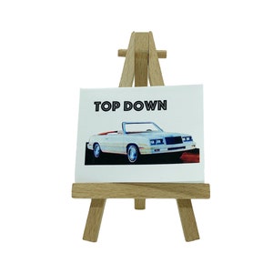 May include: A white convertible car with a red interior on a small wooden easel with the text "TOP DOWN" above the car.