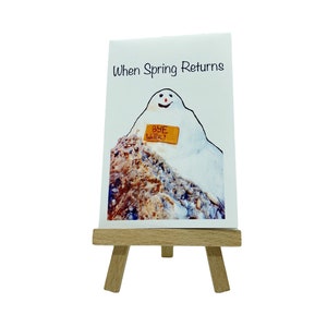 May include: A white card with a cartoon snowman melting on a rock with a sign that says "Bye Winter". The card has the text "When Spring Returns" at the top. The card is on a small wooden easel.