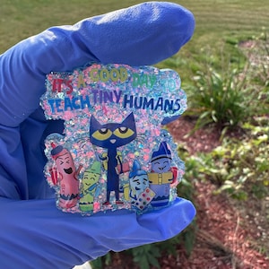 May include: A glittery, colorful resin badge with the text "IT'S A GOOD DAY TO TEACH TINY HUMANS". The badge features a cartoon cat and crayon characters. The background is a mix of pink, blue, and clear glitter.