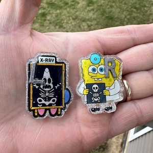 May include: Two novelty pins featuring a cartoon character. One pin is an X-ray with the text "X-RAY". The other pin is the cartoon character dressed as a doctor holding a skeleton. Both pins have glitter accents.