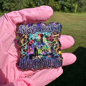 Puede incluir: Decoraci&oacute;n cuadrada con tem&aacute;tica de Hocus Pocus con el texto "It's just a bunch of Hocus Pocus". El dise&ntilde;o presenta a las hermanas Sanderson y un gato negro sobre un fondo de estampado de leopardo. Los bordes son brillantes.