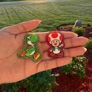May include: Two glittery resin character pins. One is green with a red and yellow belly, resembling Yoshi. The other is a mushroom-shaped character with a red and white cap, brown shoes, and a blue vest. Both pins are outlined in glitter.