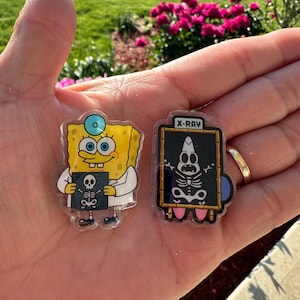 May include: Two acrylic pins featuring cartoon characters. One pin shows a yellow sponge character in a white lab coat holding an X-ray film. The second pin shows a pink starfish character behind a black board with the text X-RAY and a skeleton graphic.