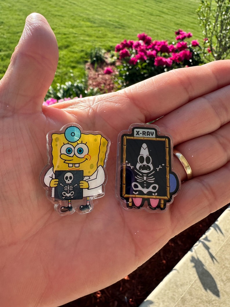 May include: Two acrylic pins featuring cartoon characters. One pin shows a yellow sponge character in a white lab coat holding an X-ray film. The second pin shows a pink starfish character behind a black board with the text X-RAY and a skeleton graphic.