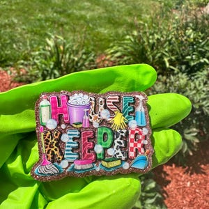 May include: A decorative badge with a glittery brown border featuring the text HOUSE KEEPER in large, colorful, patterned letters. The design includes illustrations of cleaning supplies like a mop, bucket, spray bottles, gloves, and sponges.