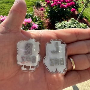 May include: Two white, semi-translucent acrylic charms with glitter accents. The left charm features the letter R above the letters BRZ. The right charm features the letter L above the letters BKS. Both charms have small white feet at the bottom.